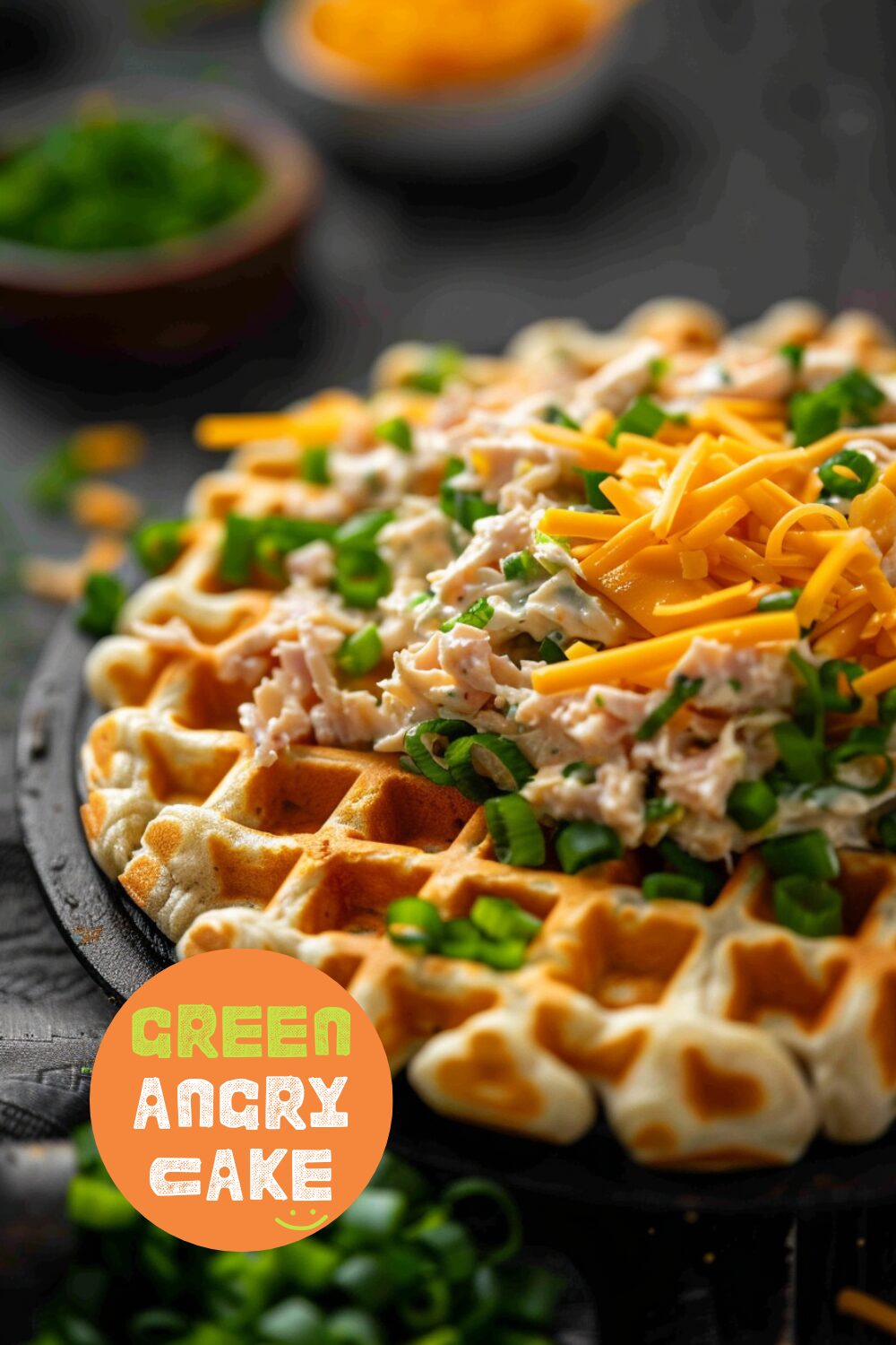 Close-up view of adding tuna salad and shredded cheddar on top of a cooked cheddar scallion waffle, ready to be baked, on a dark, black wooden texture surface.