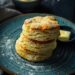 A top-down view of golden brown cheddar scallion biscuits stacked on a dark plate with a small dish of butter on a dark, black wooden texture surface.