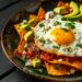 Top-down view of chilaquiles with red salsa, a sunny side up egg, avocado slices, and crumbled queso fresco on a dark wooden surface.