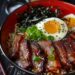 A bowl of Bacon and Egg Breakfast Ramen with thick noodles, crispy pork belly slices, a poached egg, seaweed strips, sliced green onions, katsuobushi, and toasted black sesame seeds, served in a red bowl on a dark textured surface.