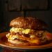 A Fairfax Breakfast Sandwich with toasted brioche bun, soft scrambled eggs, melted American cheese, caramelized onions, and sriracha mayo, served on a red plate on a dark textured surface.