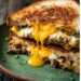 A close-up of a cheeseburger and egg grilled cheese sandwich cut in half, revealing melted cheese and juicy beef patties, on a red plate.