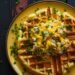 A yellow plate with a cheddar scallion waffle topped with melty cheddar cheese, tuna salad, and sliced green onions on a dark, black wooden texture surface.