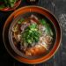 Top-down view of a bowl of pho on a blue plate, showcasing the rich broth, noodles, thinly sliced sirloin, fresh herbs, and lime wedges, all set on a dark, black wooden texture surface.