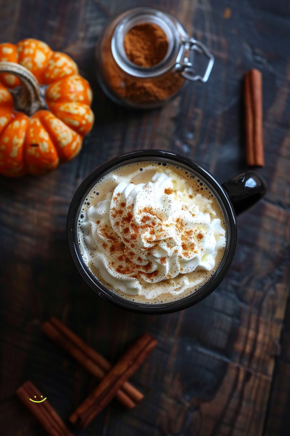 Freshly made pumpkin spice latte in a dark-colored mug, topped with whipped cream and a sprinkle of cinnamon, placed on a dark wooden surface with a small jar of pumpkin spice latte syrup beside it