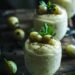 Glasses filled with gooseberry fool, topped with reserved gooseberries and decorated with mint sprigs, placed on a dark wooden surface.