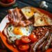 A plate of Full English breakfast with bacon, sausages, eggs, tomatoes, mushrooms, toast, and beans on a dark wooden surface.