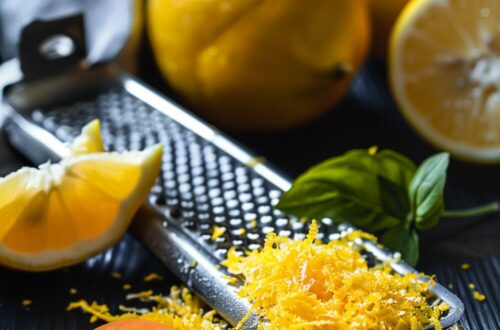 A vibrant photo showing freshly grated citrus zest in a bowl, with whole citrus fruits and a grater nearby. The background is a dark, black wooden texture surface. This image demonstrates zest preparation techniques.