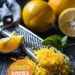 A vibrant photo showing freshly grated citrus zest in a bowl, with whole citrus fruits and a grater nearby. The background is a dark, black wooden texture surface. This image demonstrates zest preparation techniques.