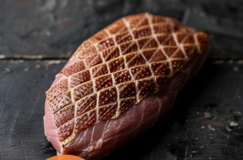 A vibrant photo showing a duck breast with perfectly scored skin in a cross-hatch pattern, ready for cooking. The background is a dark, black wooden texture surface. This image demonstrates skin scoring techniques.