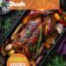 A vibrant photo showing a roasting tray with a perfectly roasted duck, golden brown and crispy, with herbs and vegetables around it. The background is a dark, black wooden texture surface. This image demonstrates duck roasting techniques.