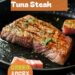 A vibrant photo showing a non-stick frying pan with a perfectly cooked tuna steak, golden brown on the outside and slightly pink in the middle, with a fish slice nearby. The background is a dark, black wooden texture surface. This image demonstrates tuna steak cooking techniques.