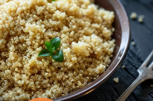 A vibrant photo showing a pan with perfectly cooked quinoa, fluffy and seasoned, with a fork nearby. The background is a dark, black wooden texture surface. This image demonstrates quinoa cooking techniques.