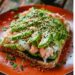 A slice of California roll avocado toast with avocado, crab, cucumber, seaweed, and sesame seeds on a [blue/orange/green/yellow/red] plate on a dark, black wooden texture surface.