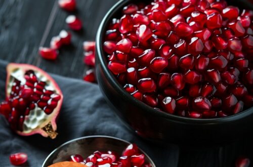 A vibrant photo showing freshly extracted pomegranate seeds in a bowl, with a halved pomegranate and a spoon nearby. The background is a dark, black wooden texture surface. This image demonstrates pomegranate preparation techniques.